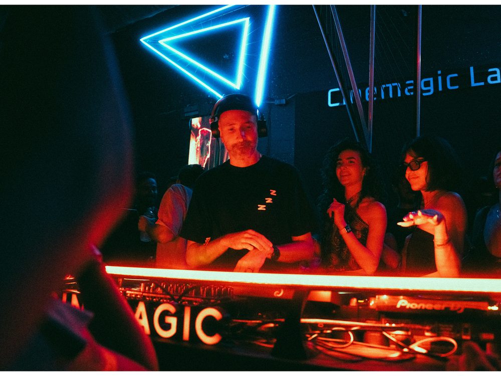 DJ and dancers at the booth beneath a blue neon triangle and the Cinemagic Lab sign